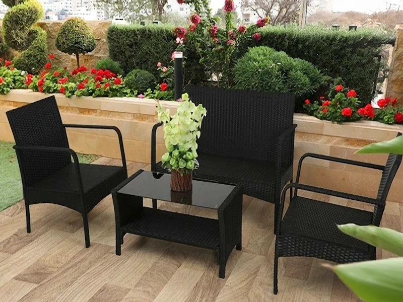 The image features a set of outdoor furniture consisting of two chairs, a sofa, and a small table, all in black wicker material. A potted plant with green leaves is placed on the table. The furniture is arranged on a tiled patio with a garden area with red flowers and dense green shrubs in the background.