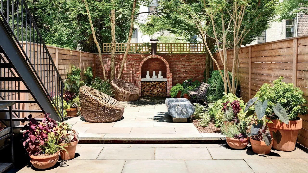 Well-maintained garden patio with terracotta pots, woven chairs, and a stone bench. Brick wall backdrop with circular opening and staircase leading to another level.