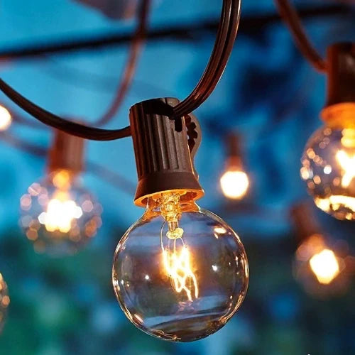 The image depicts illuminated glass bulbs hanging on black wires, set against a blurred outdoor backdrop at dusk or dawn.