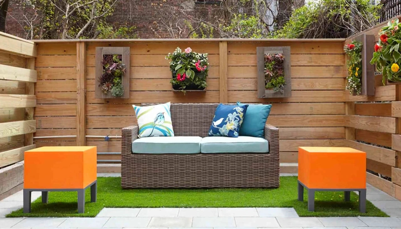 Outdoor seating area with a brown wicker sofa adorned with colorful cushions, flanked by two orange side tables on artificial grass, against a wooden slat fence with hanging planters.